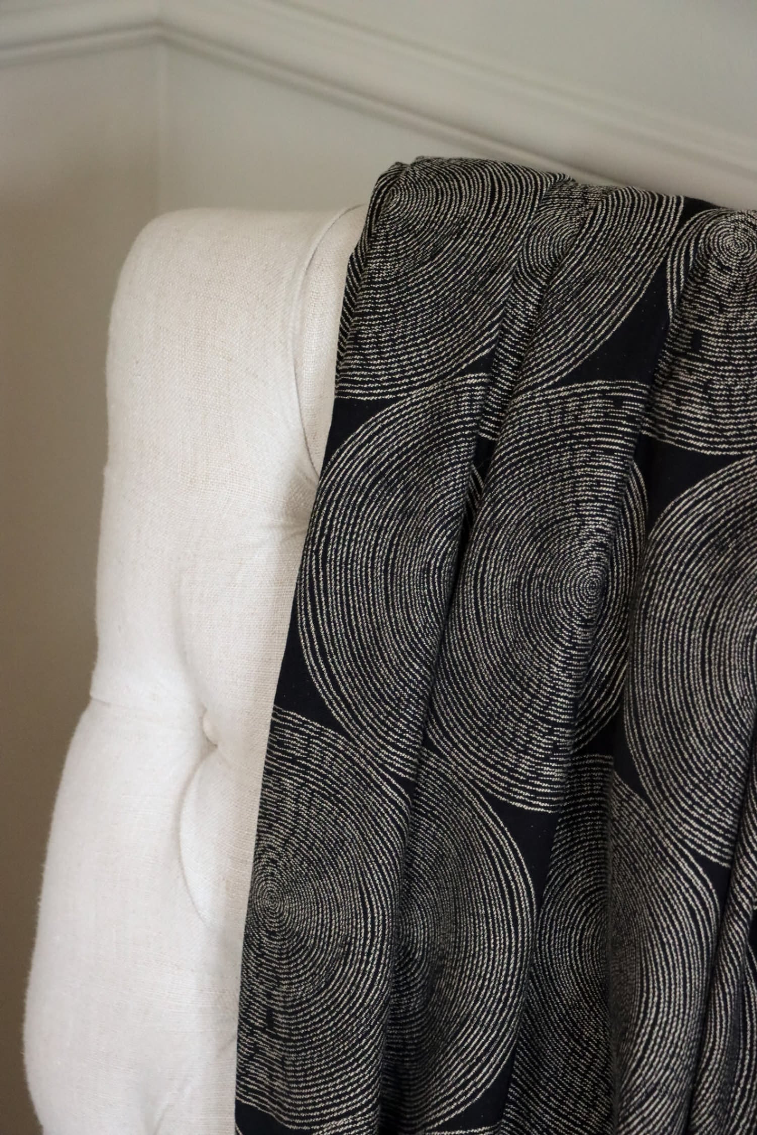 The Adam Black fabric by Arte Fresca Outlet draped loosely over a light-colored upholstered chair back, illustrating the scale of its modern concentric circle pattern and its elegant flow, perfect for assessing design impact.