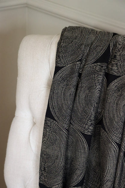The Adam Black fabric by Arte Fresca Outlet draped loosely over a light-colored upholstered chair back, illustrating the scale of its modern concentric circle pattern and its elegant flow, perfect for assessing design impact.