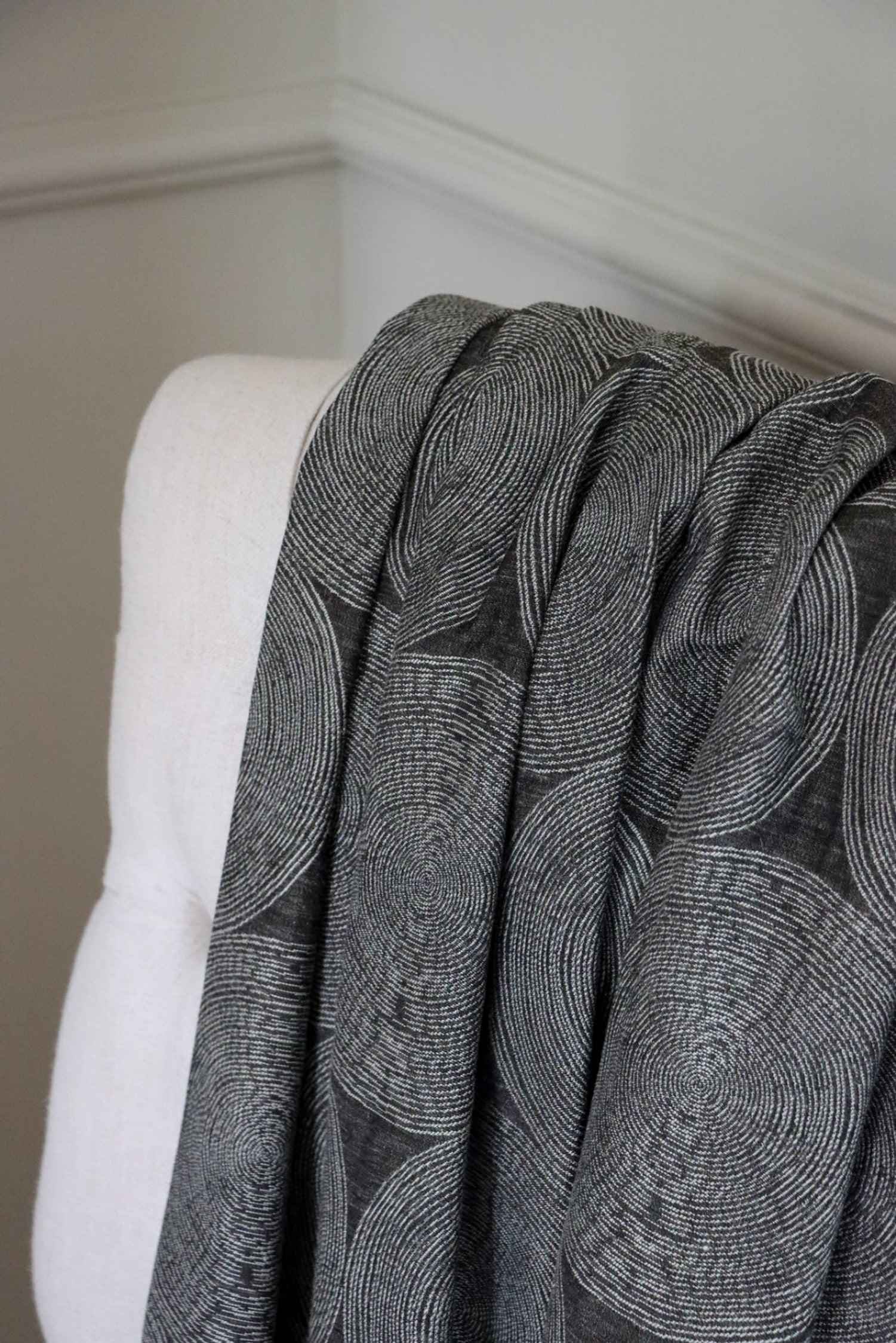 ADAM CHARCOAL fabric by Arte Fresca Outlet draped loosely over a light upholstered chair back, illustrating the scale of its concentric circle pattern in cream and silver-grey on deep anthracite, ideal for assessing design repeat and drape.
