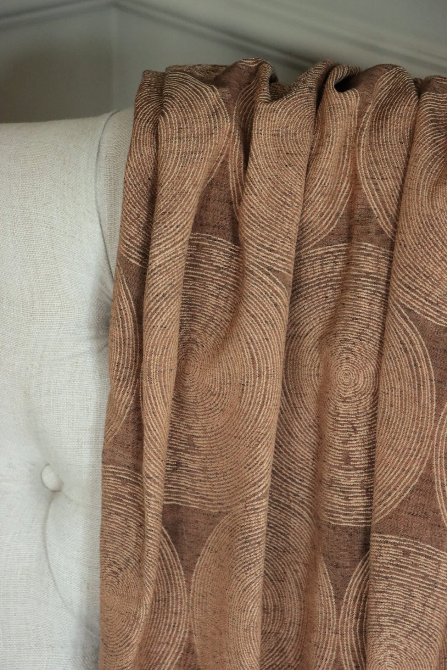 Voisin Gold fabric by Arte Fresca Outlet draped loosely over a light upholstered chair back, showcasing the scale of its subtle cream concentric circle pattern against the copper background.