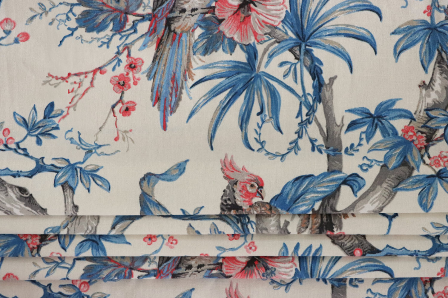 Allegria Blue/Cream fabric by Arte Fresca Outlet folded like a Roman blind, illustrating its structured appearance and the exotic tropical design with the parrot and hibiscus pattern, perfect for window treatments.


