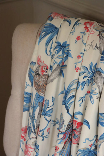 Allegria Blue/Cream fabric by Arte Fresca Outlet draped loosely over a light upholstered chair back, showcasing the scale of its tropical design with the parrot and hibiscus pattern in blue, red, and beige on cream.