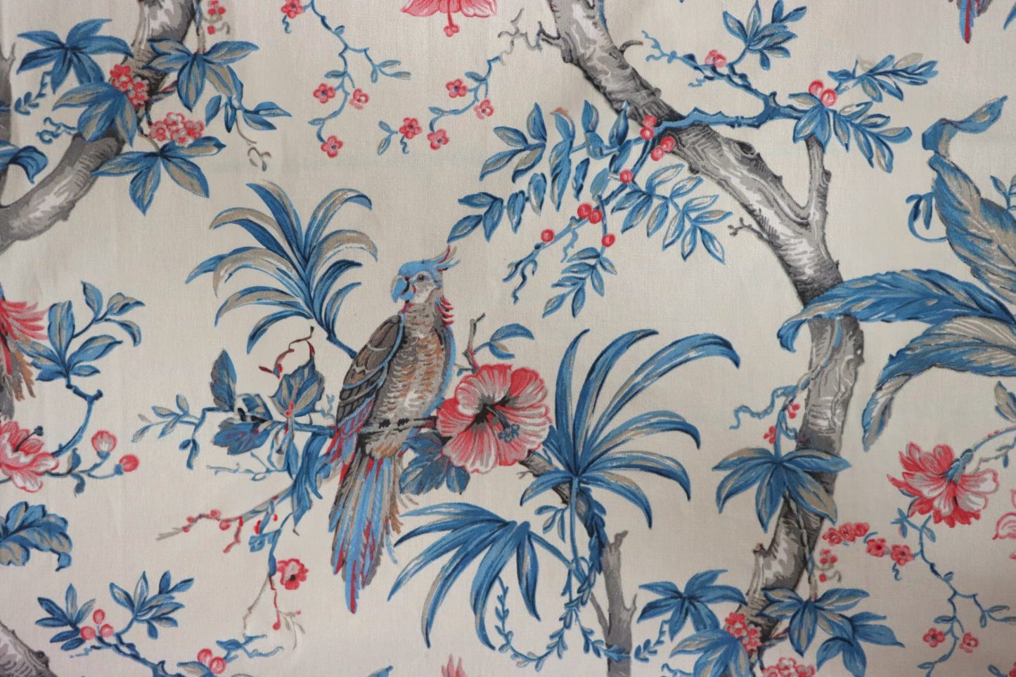 Allegria Blue/Cream fabric by Arte Fresca Outlet, showing a large section of the woven cotton fabric with a detailed tropical design featuring a vibrant parrot, hibiscus flowers, and foliage in blue, red, and beige on a cream background.