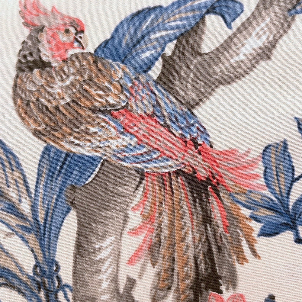 Close-up of ALLEGRIA BLUE/CREAM fabric coupon from Arte Fresca Outlet, showcasing the intricate details of the tropical pattern. A colorful parrot, hibiscus flowers, and leaves are depicted with fine lines and careful drawing in blue, red, and beige on a cream 100% cotton textile, highlighting its exquisite quality.