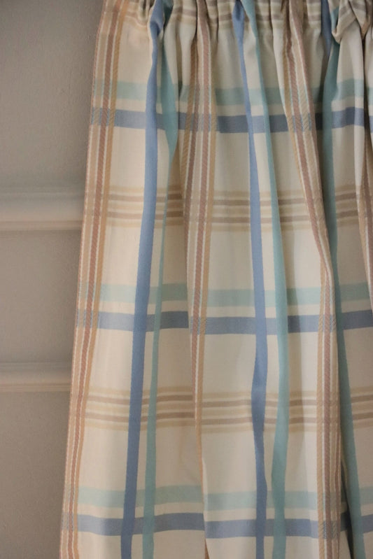 BRAXTON CHECK BLUE/CREAM fabric from Arte Fresca Outlet, elegantly folded in drapes like a curtain, showcasing its soft fall, the harmonious blue, cream, and brown check pattern, and its suitability for stylish window treatments.