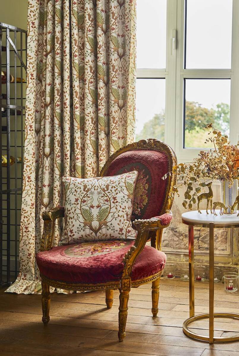 A vintage-style upholstered armchair with a deep red velvet seat and back, gold carved wood arms and legs, and a matching floral patterned throw pillow, set in a sunlit room next to a window and a small round side table.