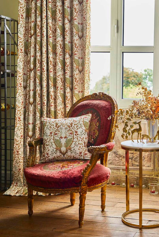 A vintage-style upholstered armchair with a deep red velvet seat and back, gold carved wood arms and legs, and a matching floral patterned throw pillow, set in a sunlit room next to a window and a small round side table.