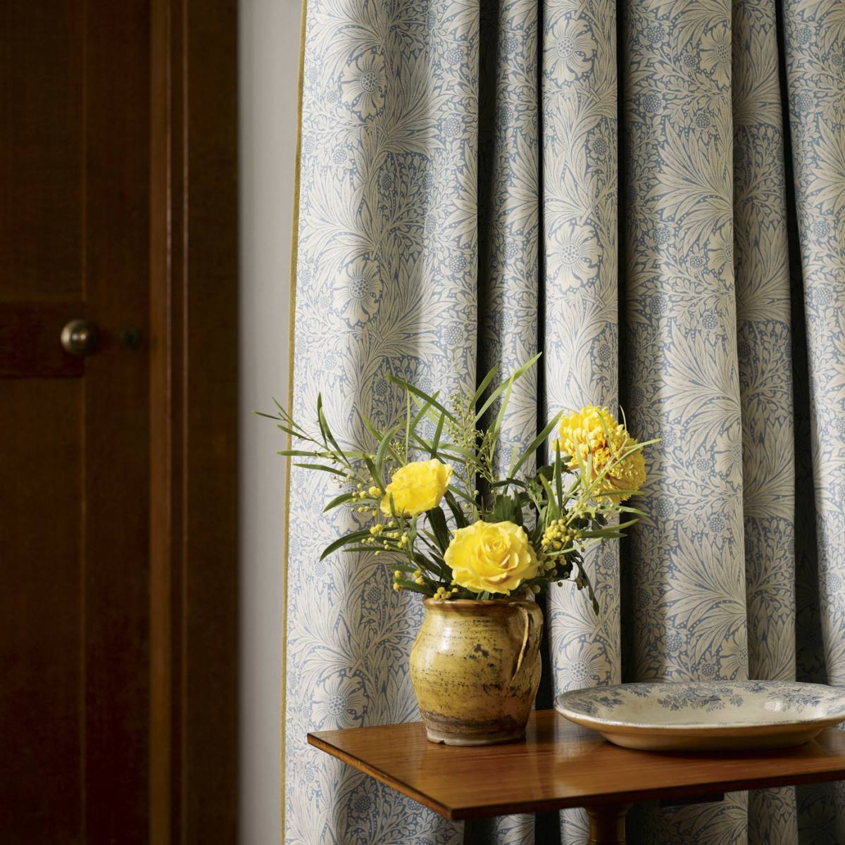 Blue and white patterned curtains hanging on a window next to a dark wooden door, with a small wooden table holding a vase of yellow flowers and a decorative plate in the foreground.