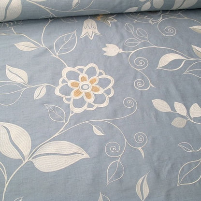 Close-up of MOREEN AQUA embroidered silk fabric from Arte Fresca Outlet, revealing the raised, intricate floral embroidery. Silvery ivory and creamy beige threads form decorative blossoms and swirling stems on the smooth, shimmering aqua light-blue silk-rayon ground, showcasing its delicate artistry and luxurious texture.