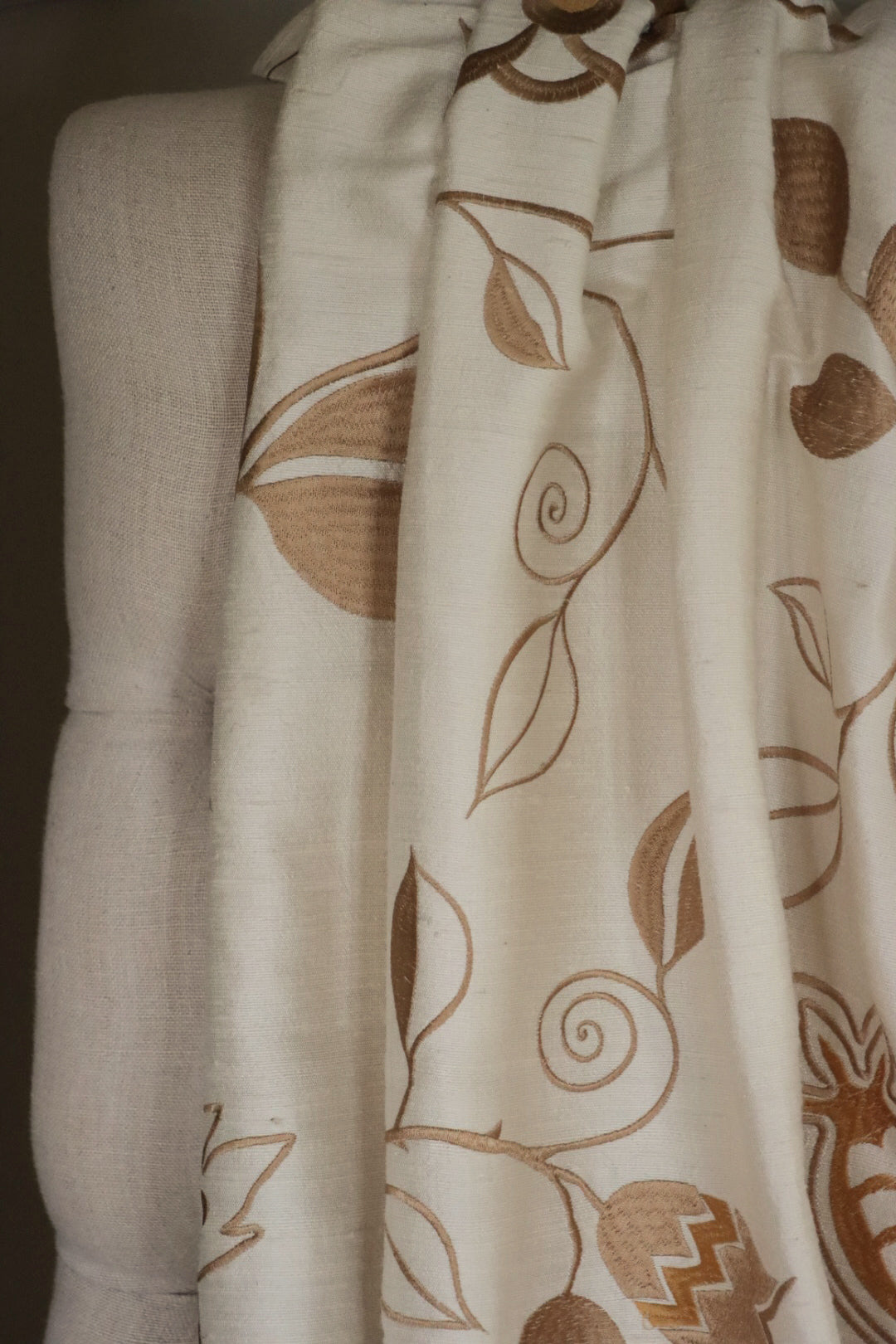 MOREEN CAMEL silk fabric by Arte Fresca Outlet draped loosely over a light upholstered chair back, illustrating the elegant scale of its embroidered floral pattern in brown on camel, ideal for assessing design repeat and luxurious drape for home decor.
