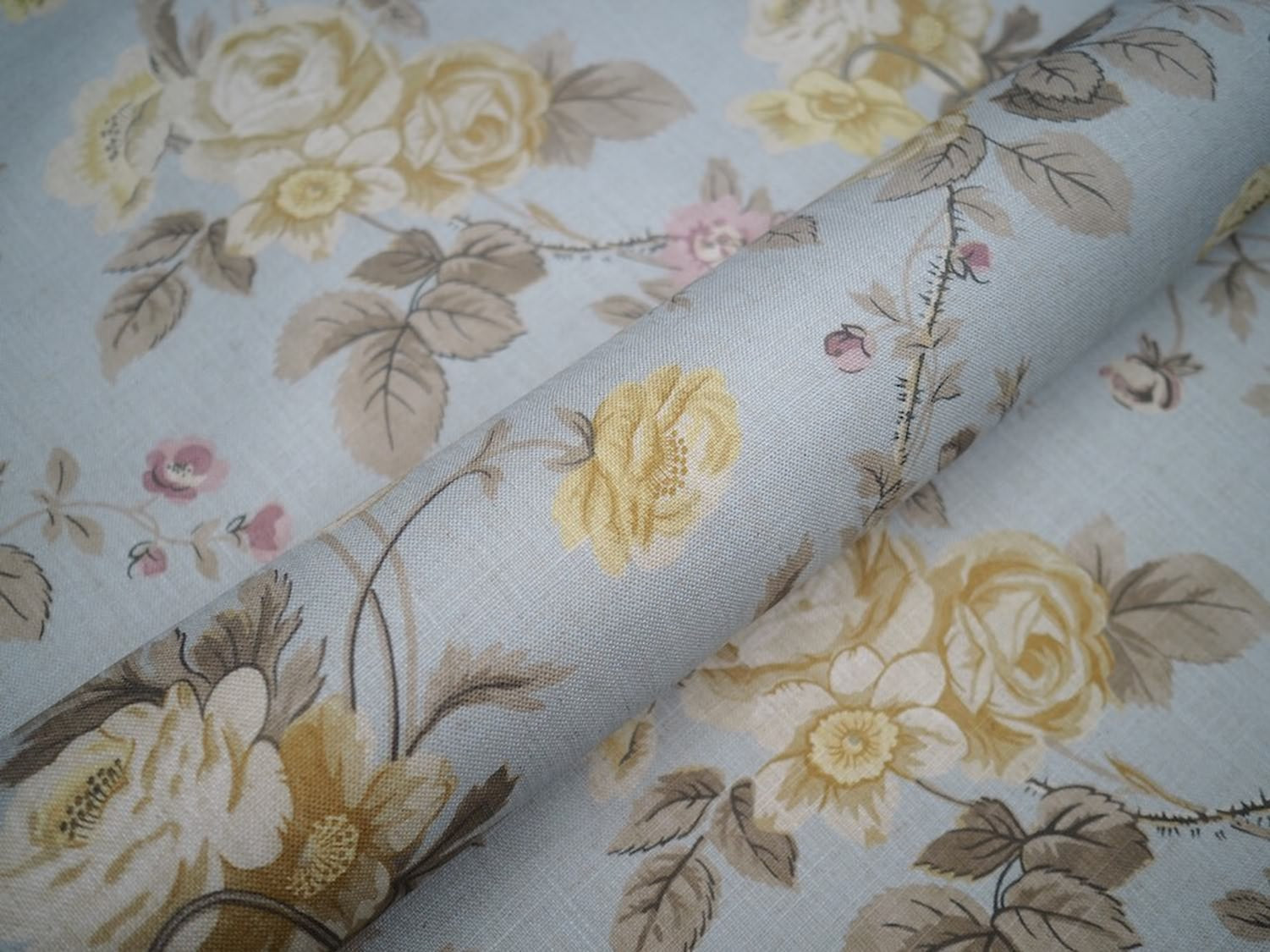 Close-up of PHILIPPA PALE BLUE fabric from Arte Fresca Outlet, revealing the detailed rose, blossom, and leaf motifs, soft cream, pink, and muted grey tones on a pale blue linen blend with subtle woven texture.