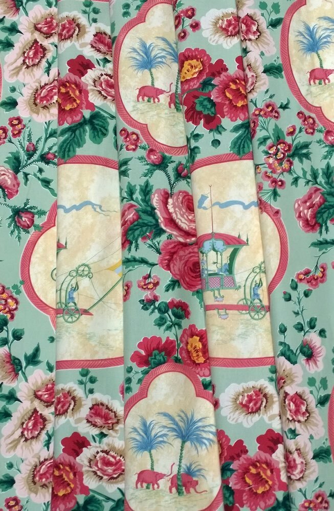 A close-up of a fabric or wallpaper pattern featuring a toile-style design with pink and red flowers, green leaves, and cream cartouches containing palm trees and small animal motifs on a pale mint background.
