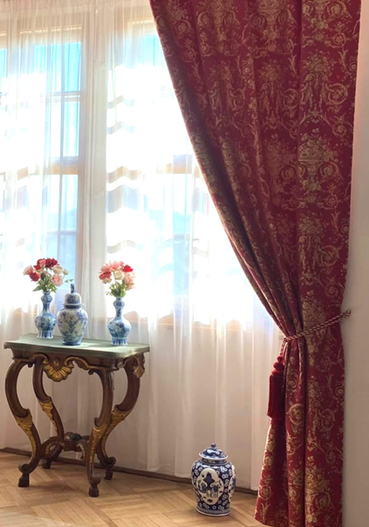 The VILLA BORGHESE fabric in a red and gold colorway, used as a luxurious window curtain. The image demonstrates the fabric's heavy drape and how its opulent pattern adds a stately and elegant feel to a room.