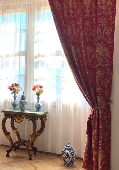 The VILLA BORGHESE fabric in a red and gold colorway, used as a luxurious window curtain. The image demonstrates the fabric's heavy drape and how its opulent pattern adds a stately and elegant feel to a room.