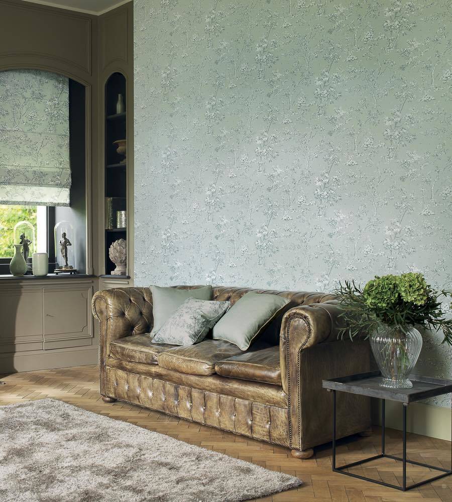 A styled living room featuring a distressed brown leather Chesterfield sofa with matching throw pillows, set against a wall with textured sage green wallpaper. A small side table with a green plant sits to the right, and a gray shag rug is on the floor.